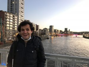 Faruk is standing on the bridge above the Meuse river, smiling at the camera.