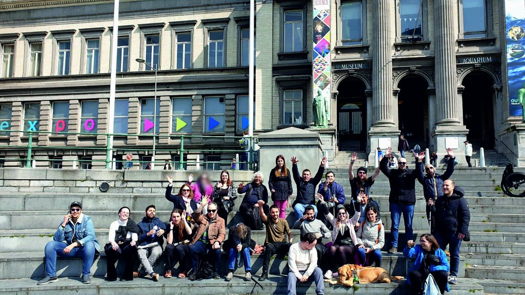 the group of participants sit on the stairs in front of the Aquarium Museum while waving and smiling at the camera