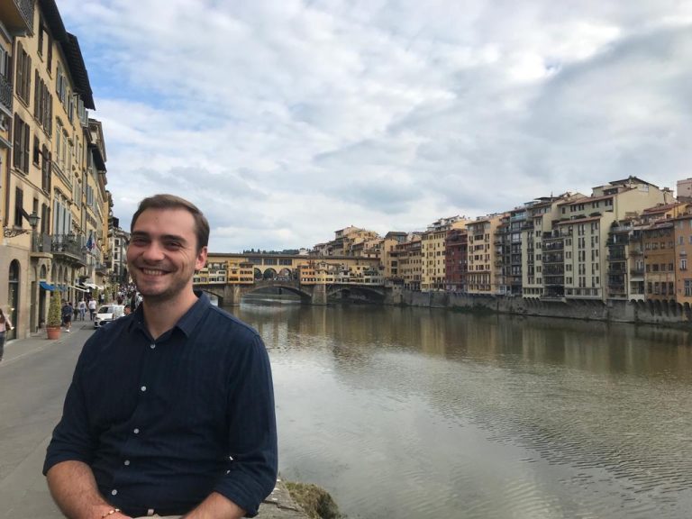 Mihai is sitting while smiling at the camera. In the background is a river with houses on both sides of it.