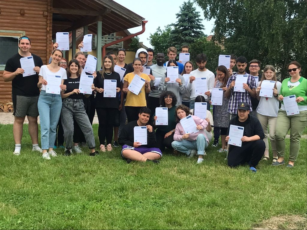 Group of the participants in Youth Exchange take a picture in the grass, while showing their Youthpass certificates