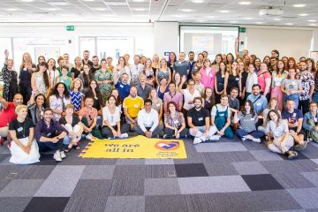 Group photo of many people gathered around a sign saying "we are all in"