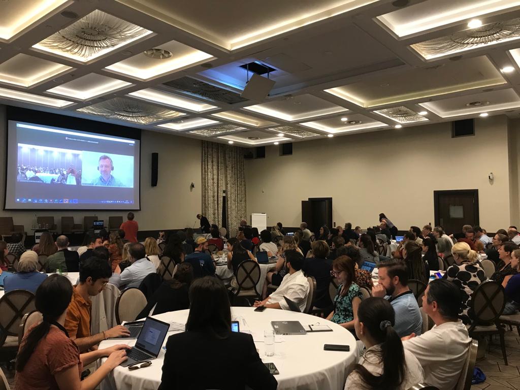 People in a room listening to the conference, a speaker is presenting in videoconference.