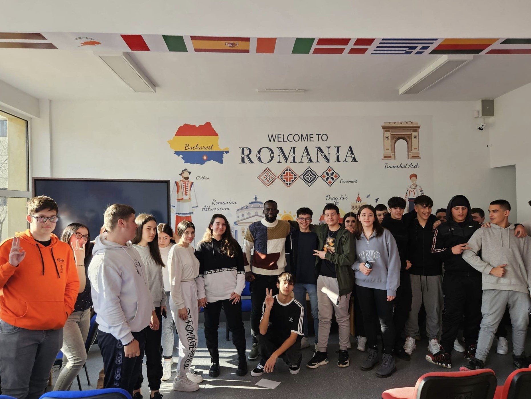 Anca, Abraham, the SAKURA NGO partners and numerous young volunteers pose all together in a room full of flags of European countries. On the wall behind them, the writing "Welcome to Romania"