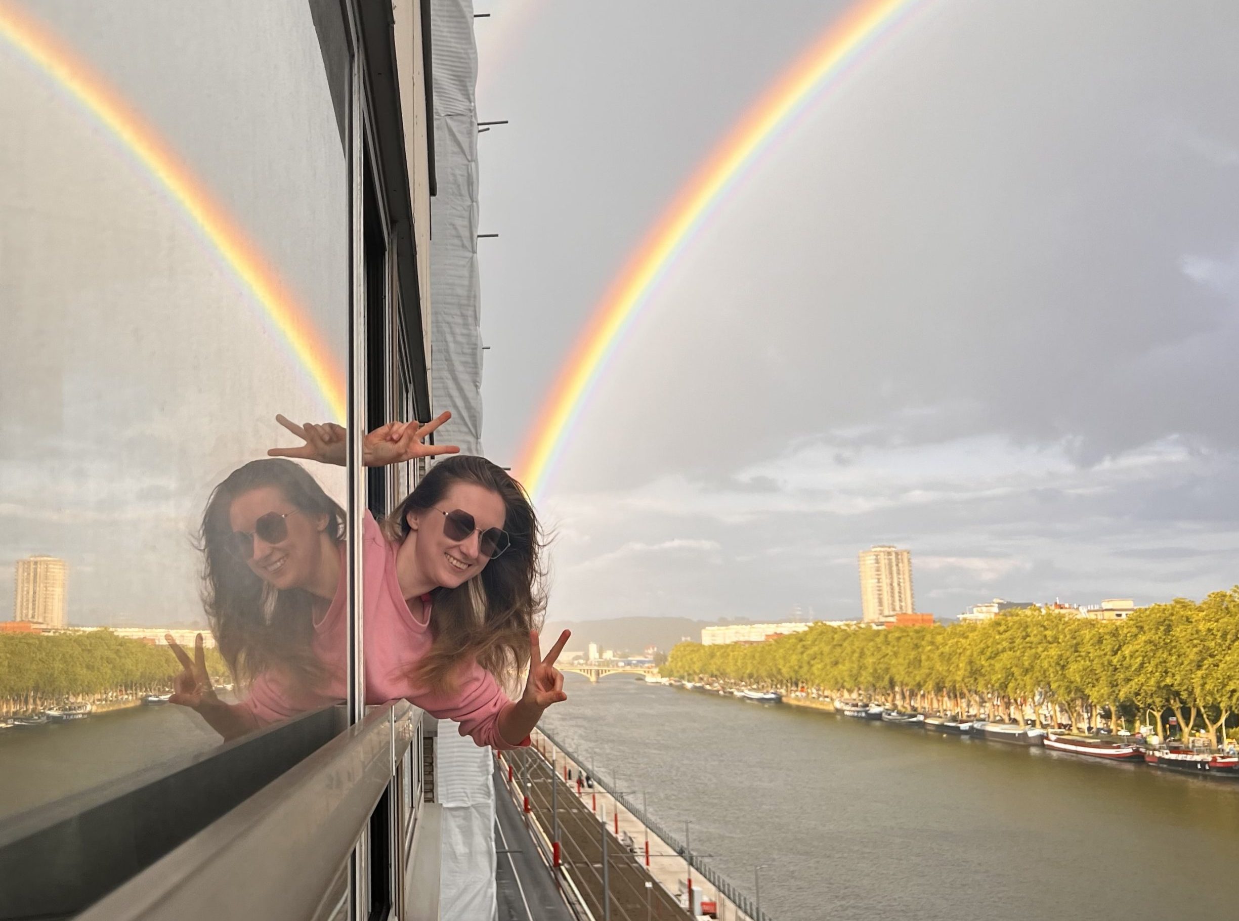 Portait picture of a smiling Kinga peeking outside of a ferry window with two rainbows in the background.