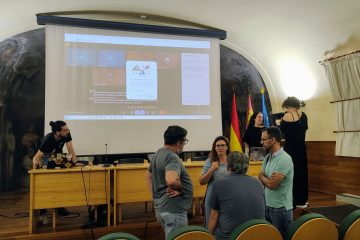 Some event participants in the room before the event, discussing. On the podium, the Arkhe organisers are setting up the Zoom meeting for online participants, while talking to one of the TBT training course participants from Spain.