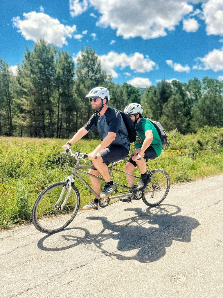 Participants wearing helmets riding a tandem bicycle along a sunlit country road with a green landscape behind them.