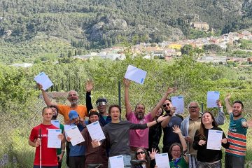 Participants standing outdoors holding up white certificates, with a tandem bicycle resting in front of them against a mountain backdrop.