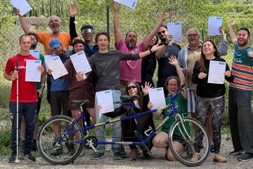 Participants standing outdoors holding up white certificates, with a tandem bicycle resting in front of them against a mountain backdrop.