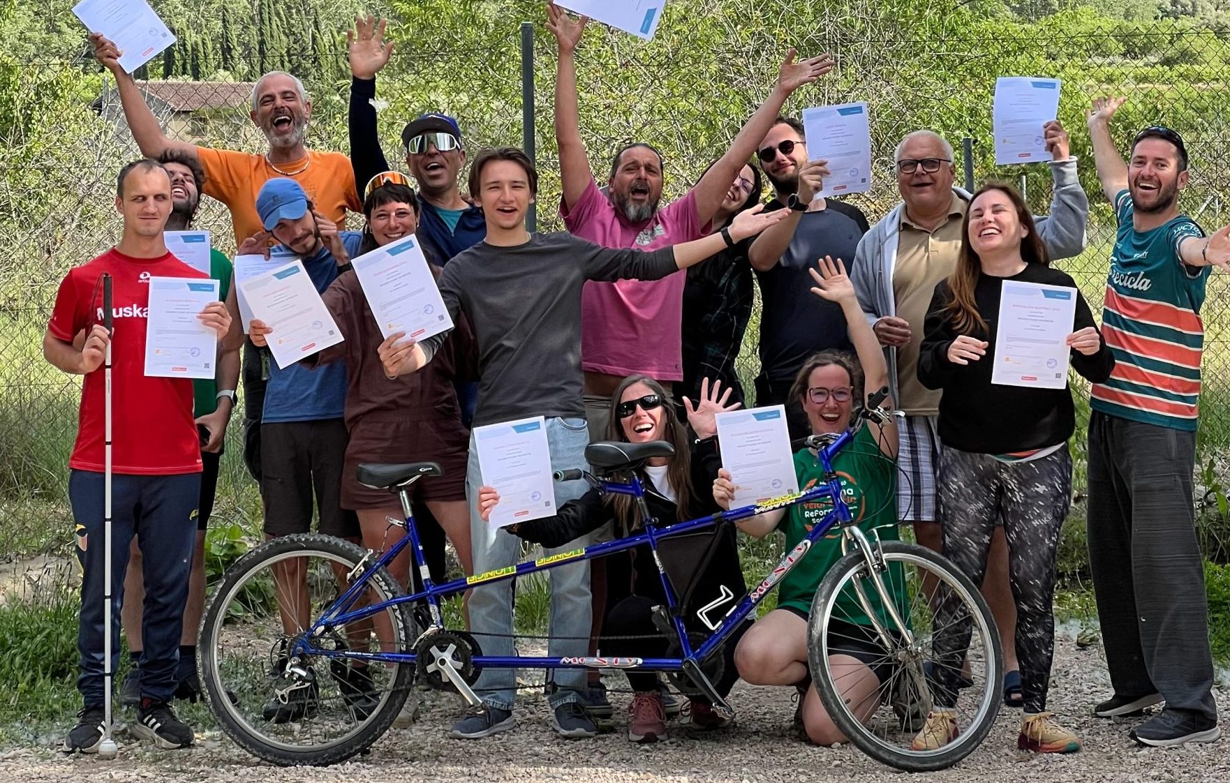 Participants standing outdoors holding up white certificates, with a tandem bicycle resting in front of them against a mountain backdrop.