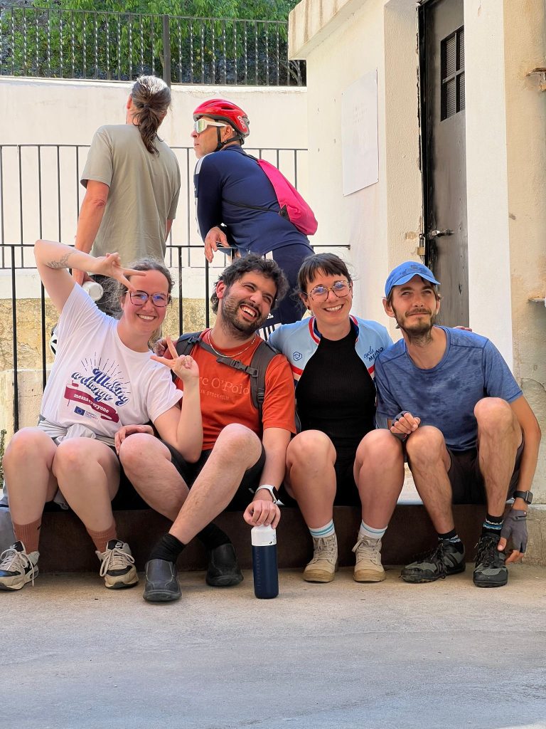 Youssri and Zoe with two other participants sitting side-by-side on a low concrete step, smiling warmly at the camera while wearing casual athletic gear.