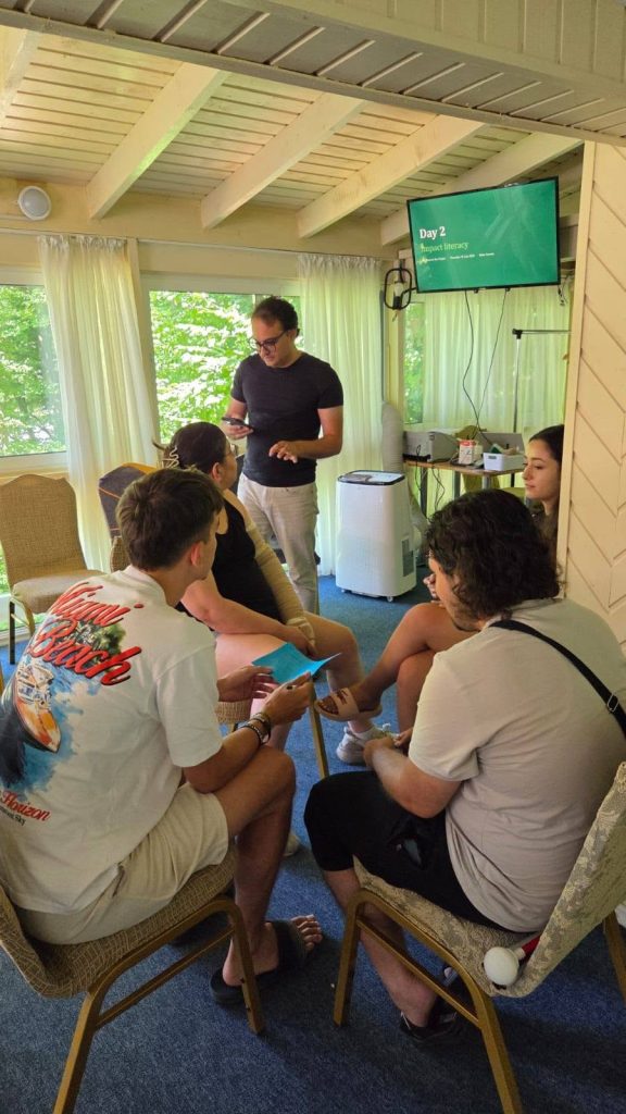 Participants take part in a small group discussion during the Erasmus+ Youth Seminar in Romania. A facilitator stands in the centre guiding the activity, while a presentation on "Impact Literacy" is displayed on a screen in the background.