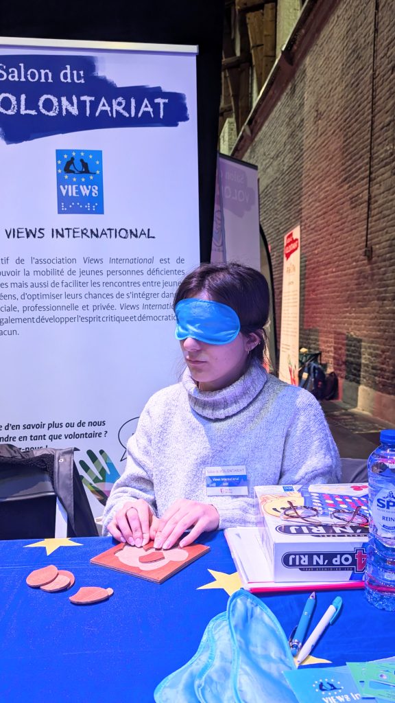 Sofia blindfolded completing a puzzle at the Volunteering Fair in Liège. Behind her, a roll-up presenting VIEWS International