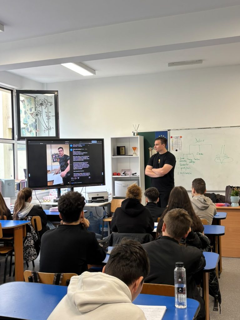 Silviu stands beside a screen showing a social media post about his volunteering activities, while students seated at their desks follow the presentation.