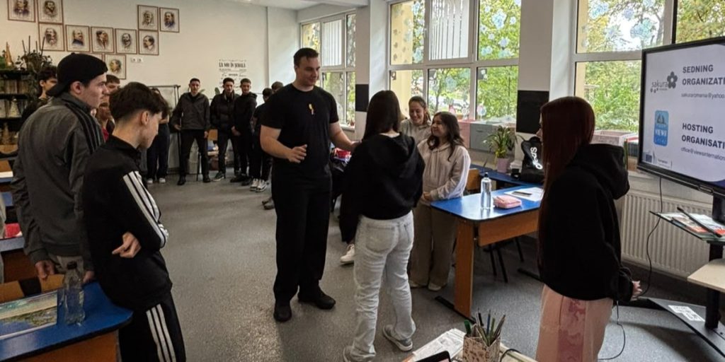 Silviu stands in a classroom surrounded by students taking part in a paired guiding activity. Several students watch as two participants stand in the centre of the room.