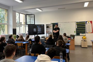 Silviu stands at the front of a classroom, presenting his European Solidarity Corps volunteering experience to students seated at their desks. A large screen displays content from VIEWS International.