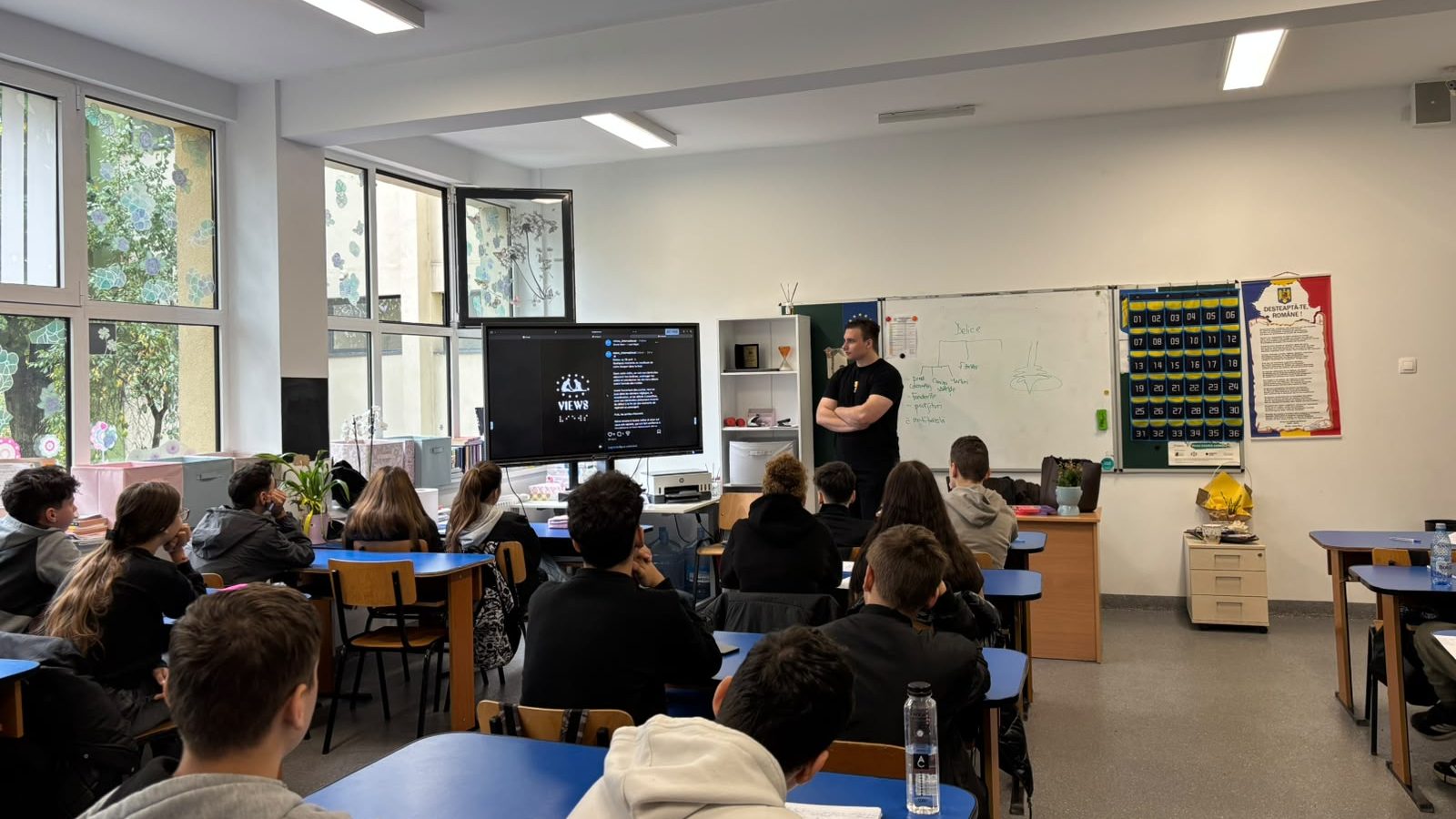 Silviu stands at the front of a classroom, presenting his European Solidarity Corps volunteering experience to students seated at their desks. A large screen displays content from VIEWS International.